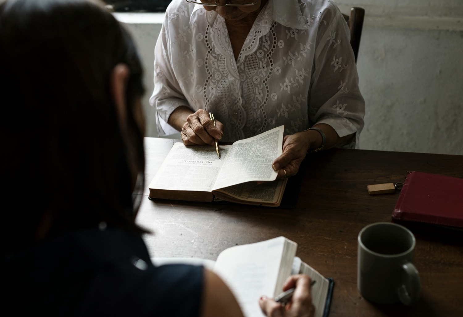An older woman studying an open Bible with a younger woman taking notes